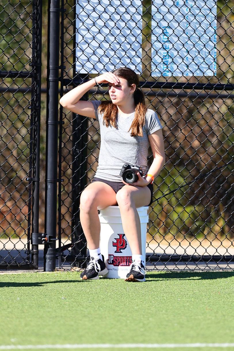 A parent with a camera sitting on a bucket just inside the field fence